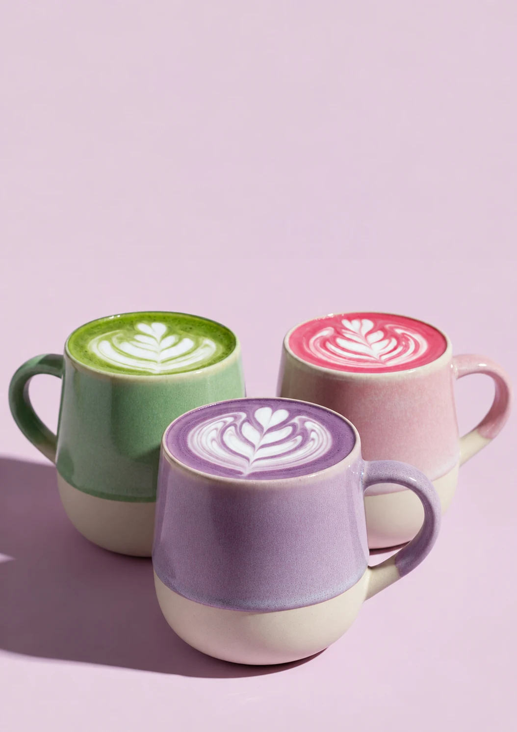 Tre koppar med gröna, rosa och lila lattedrycker.